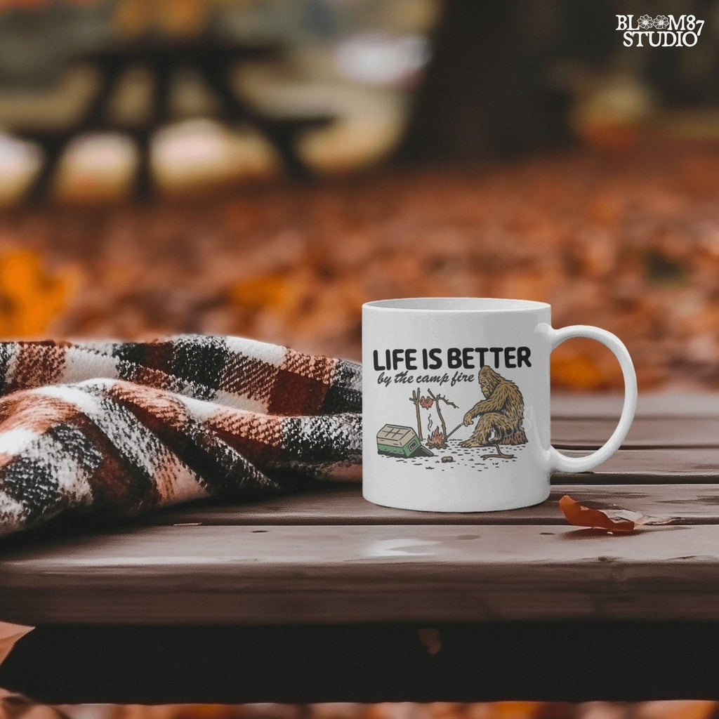 Bigfoot roasting meat over a campfire next to a tipped cooler, with the phrase “Life Is Better by the camp fire” above, illustrated PNG design for outdoor sublimation.