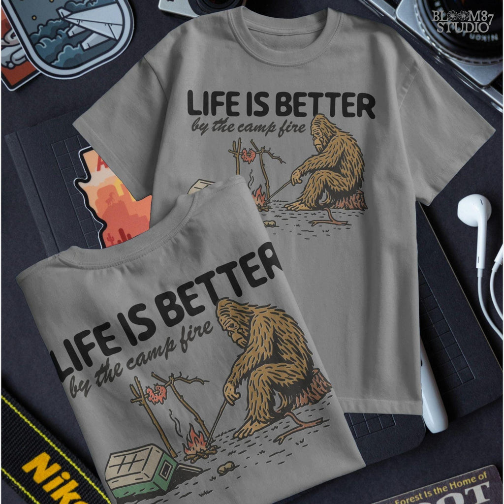 Bigfoot roasting meat over a campfire next to a tipped cooler, with the phrase “Life Is Better by the camp fire” above, illustrated PNG design for outdoor sublimation.