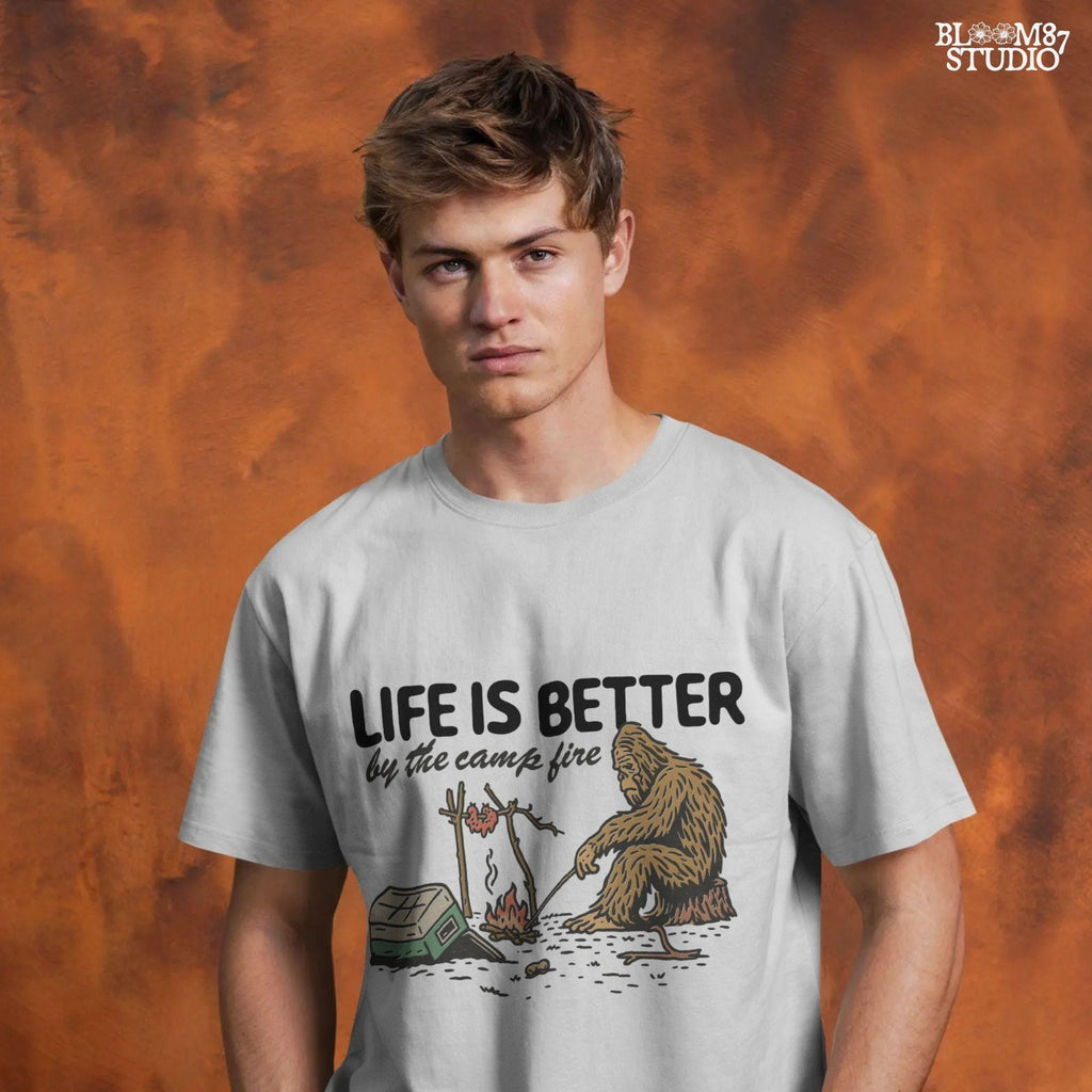 Bigfoot roasting meat over a campfire next to a tipped cooler, with the phrase “Life Is Better by the camp fire” above, illustrated PNG design for outdoor sublimation.