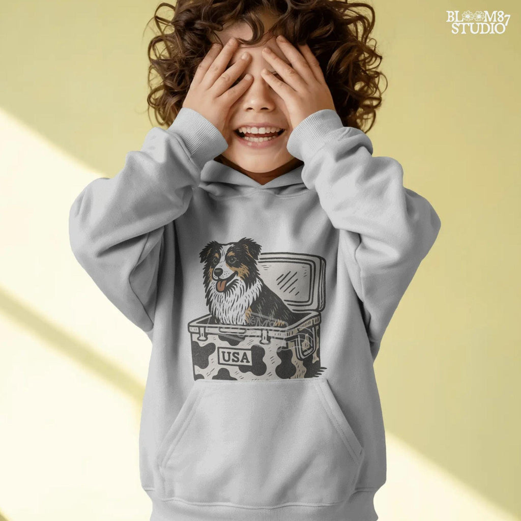 Illustration of an Australian Shepherd dog sitting inside a cowhide-pattern USA cooler; western patriotic sublimation PNG for dog lovers and crafters.