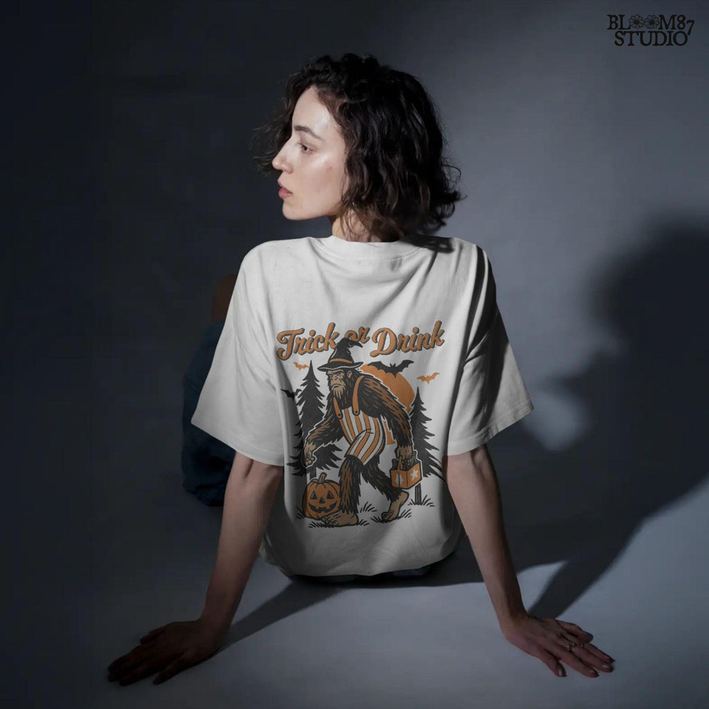 Funny Halloween bigfoot holding beer in witch hat and striped overalls, walking past pumpkins and trees with “Trick or Drink” text above.