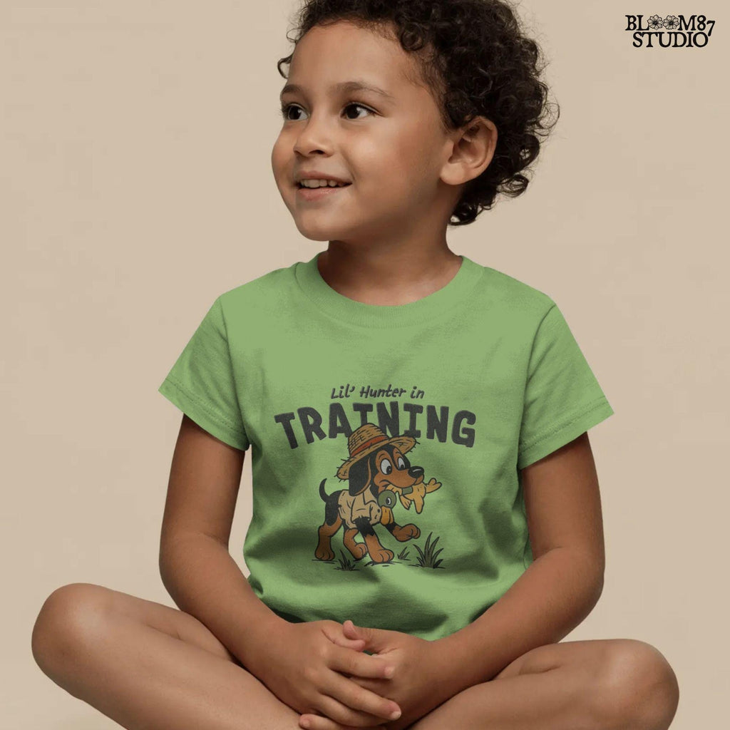 Cartoon hunting dog wearing straw hat and camo shirt holding duck toy in mouth with “Lil’ Hunter in Training” text – cute PNG for kids sublimation.