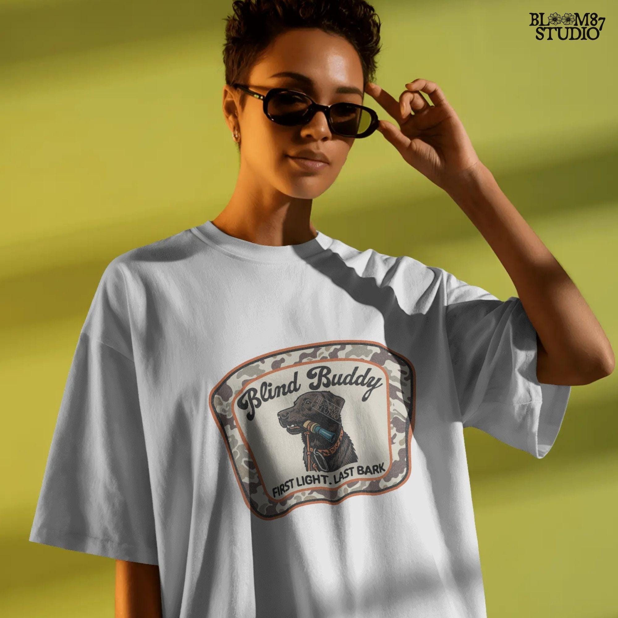 Black labrador dog holding a duck call in its mouth, centered in a camouflage hunting patch with text “Blind Buddy – First Light. Last Bark”