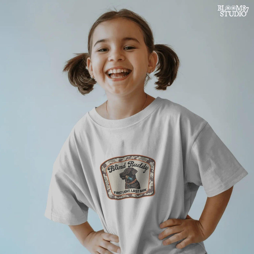 Black labrador dog holding a duck call in its mouth, centered in a camouflage hunting patch with text “Blind Buddy – First Light. Last Bark”