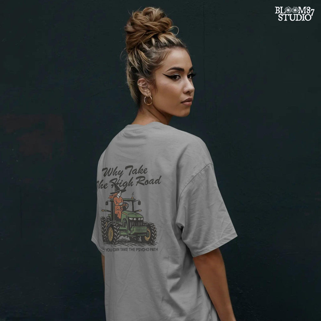 Funny illustration of a red-haired witch in a black hat driving a green tractor with the quote “Why take the high road when you can take the psycho path,” in rustic cartoon style.