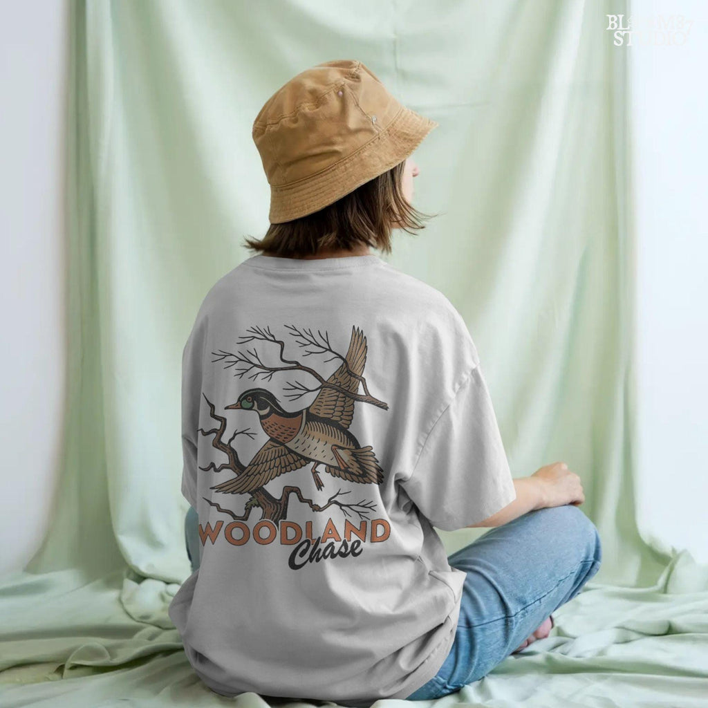 Vintage wood duck flying over bare branches with “Woodland Chase” text in rustic orange and black; sublimation PNG for hunting or fall apparel.