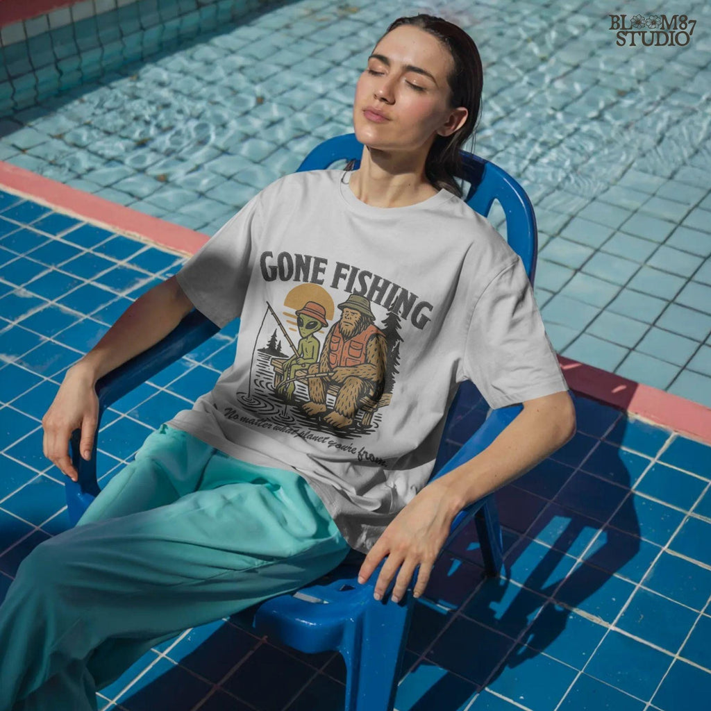 Funny illustration of Bigfoot and an alien fishing together on a lake dock at sunset, wearing fishing hats and vests—perfect for summer and Father’s Day sublimation.