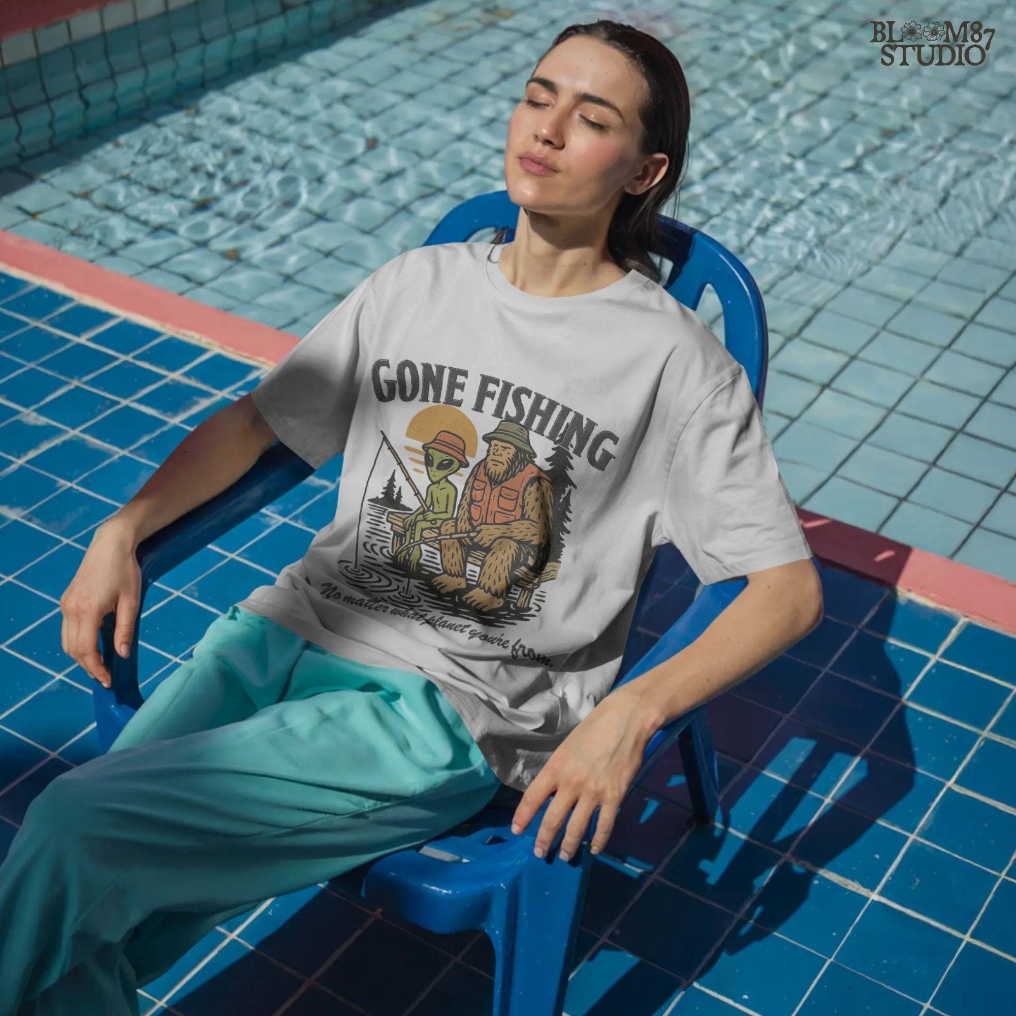 Funny illustration of Bigfoot and an alien fishing together on a lake dock at sunset, wearing fishing hats and vests—perfect for summer and Father’s Day sublimation.