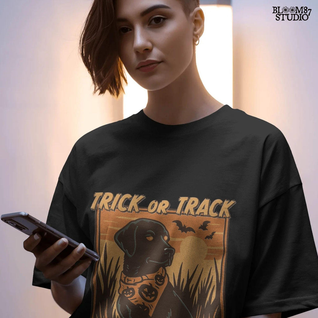 Halloween PNG of a black lab hunting dog wearing a pumpkin bandana with bats, sunset, and text “Trick or Track – He Hunts More Than Candy”