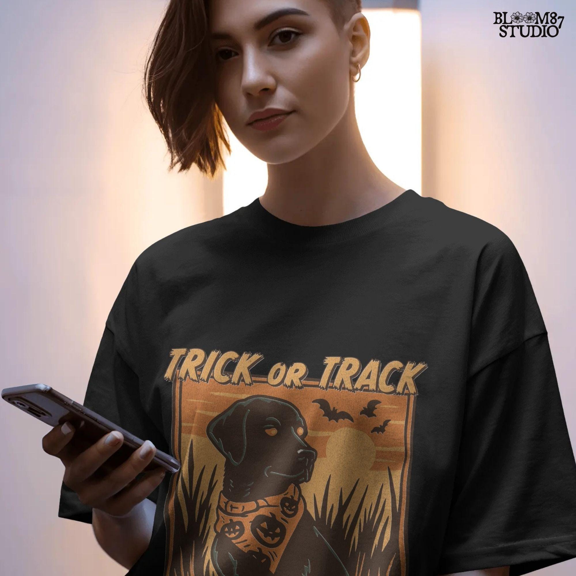 Halloween PNG of a black lab hunting dog wearing a pumpkin bandana with bats, sunset, and text “Trick or Track – He Hunts More Than Candy”