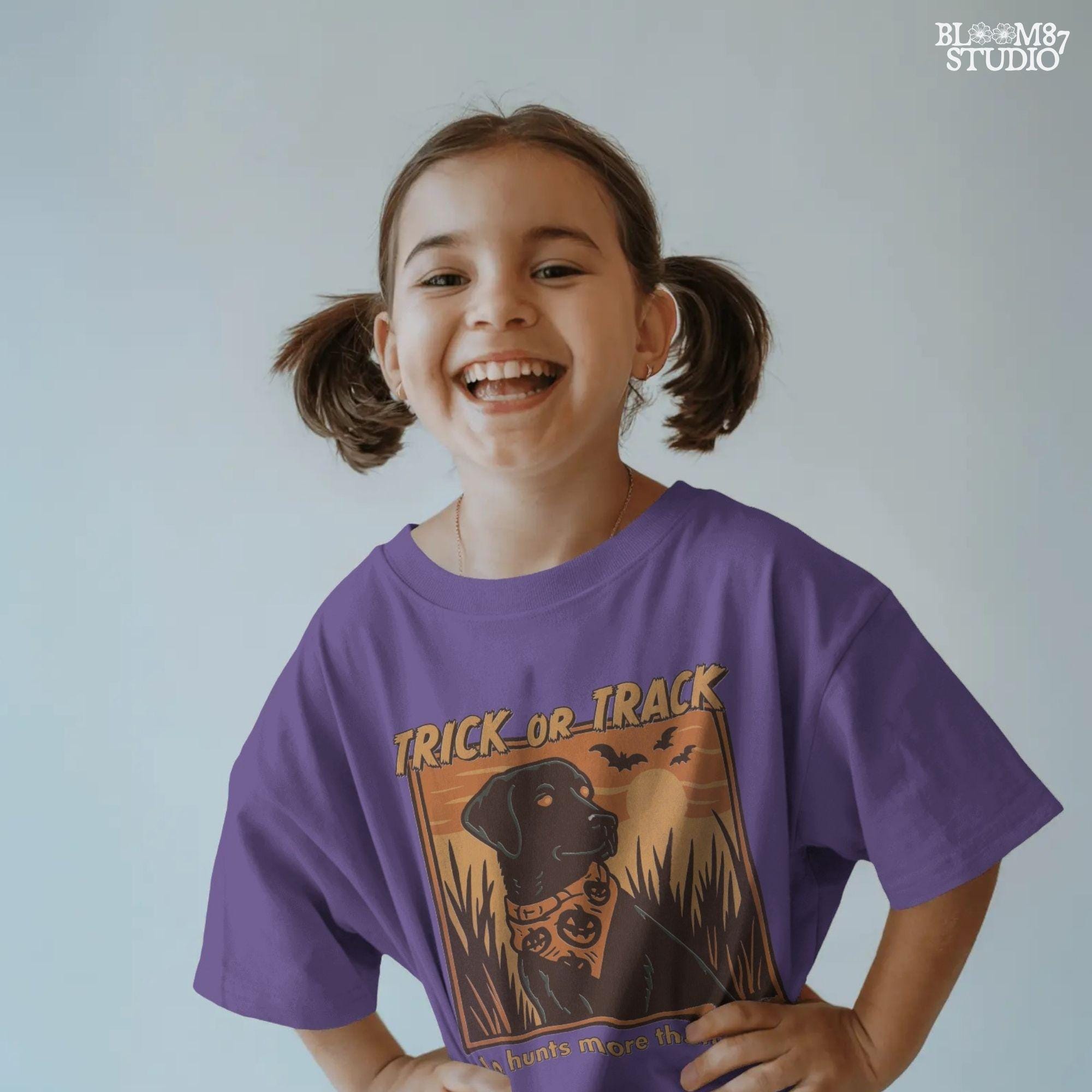 Halloween PNG of a black lab hunting dog wearing a pumpkin bandana with bats, sunset, and text “Trick or Track – He Hunts More Than Candy”