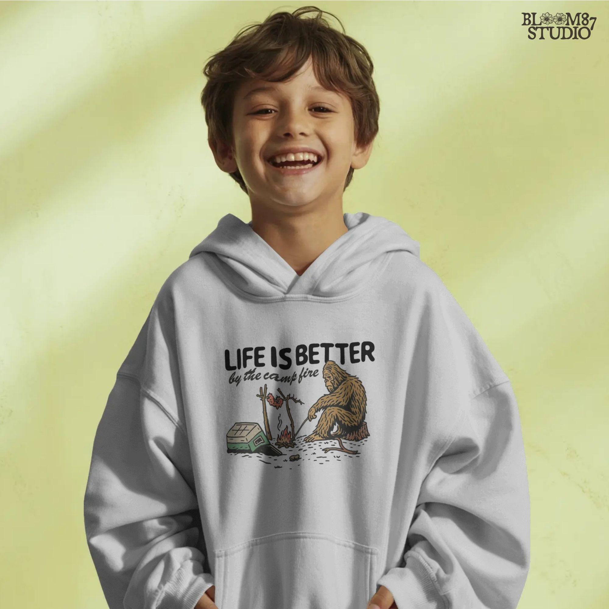 Bigfoot roasting meat over a campfire next to a tipped cooler, with the phrase “Life Is Better by the camp fire” above, illustrated PNG design for outdoor sublimation.