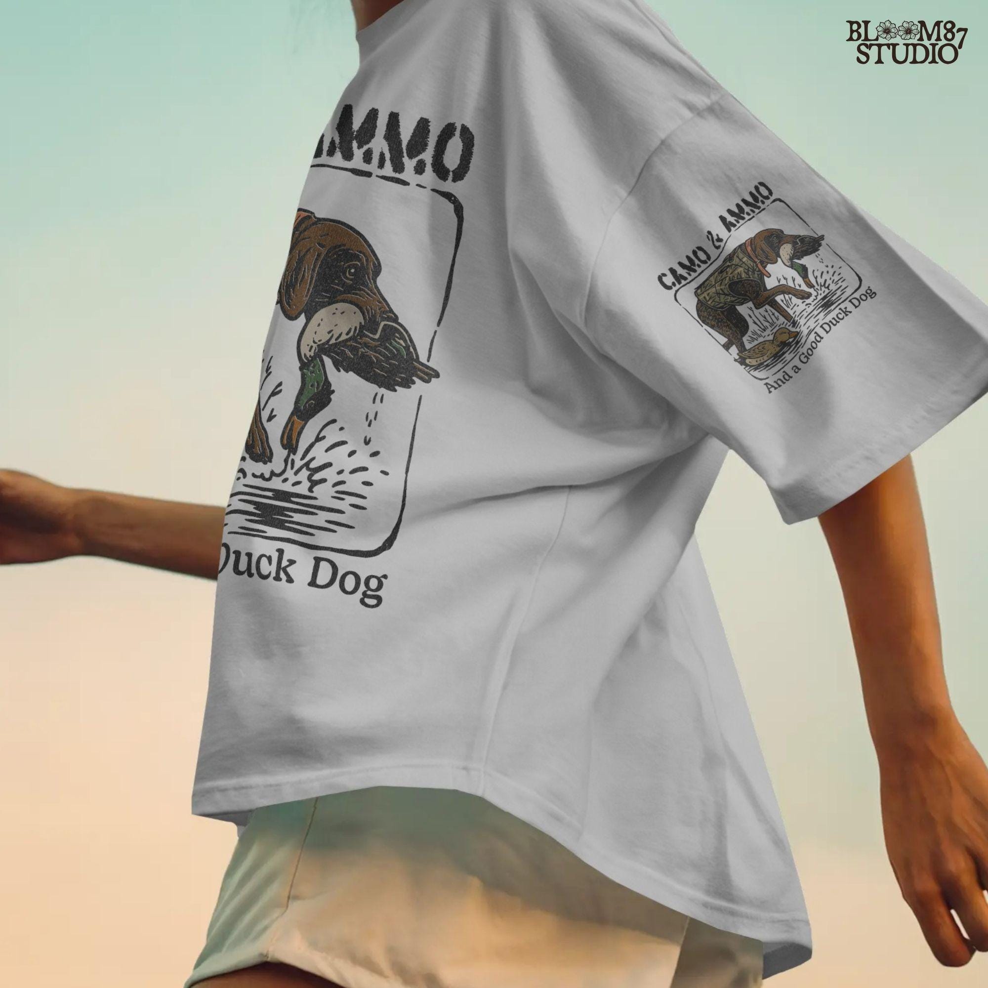 Hunting dog in a camo vest retrieving a mallard duck with a swimming duck nearby and text saying “Camo & Ammo and a Good Duck Dog”.