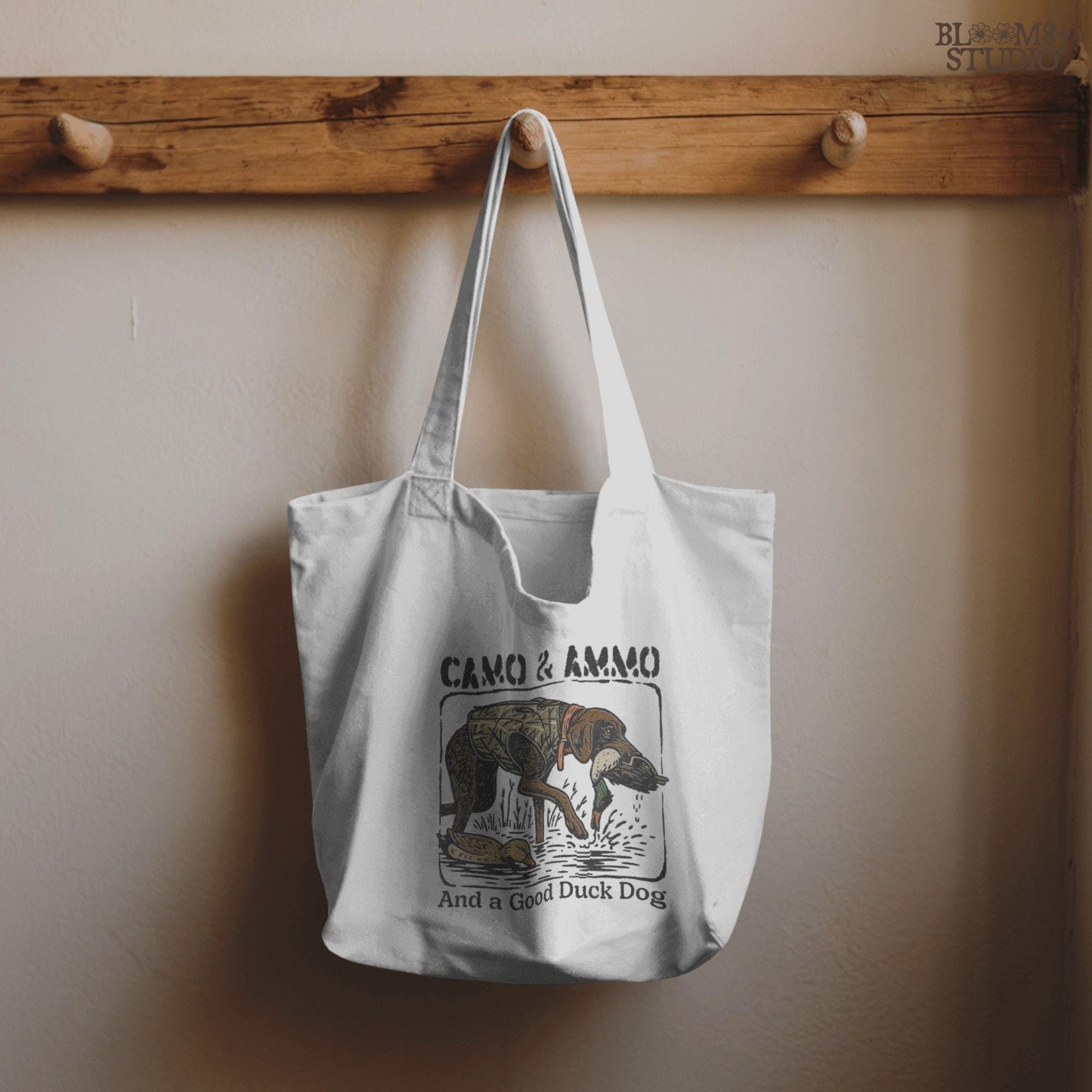 Hunting dog in a camo vest retrieving a mallard duck with a swimming duck nearby and text saying “Camo & Ammo and a Good Duck Dog”.