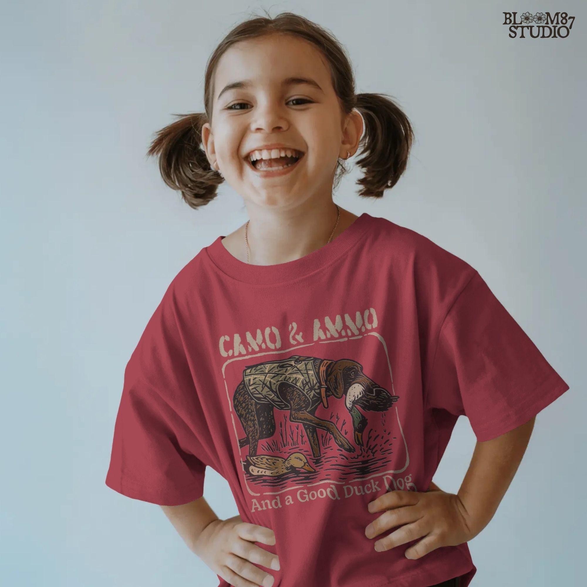 Hunting dog in a camo vest retrieving a mallard duck with a swimming duck nearby and text saying “Camo & Ammo and a Good Duck Dog”.