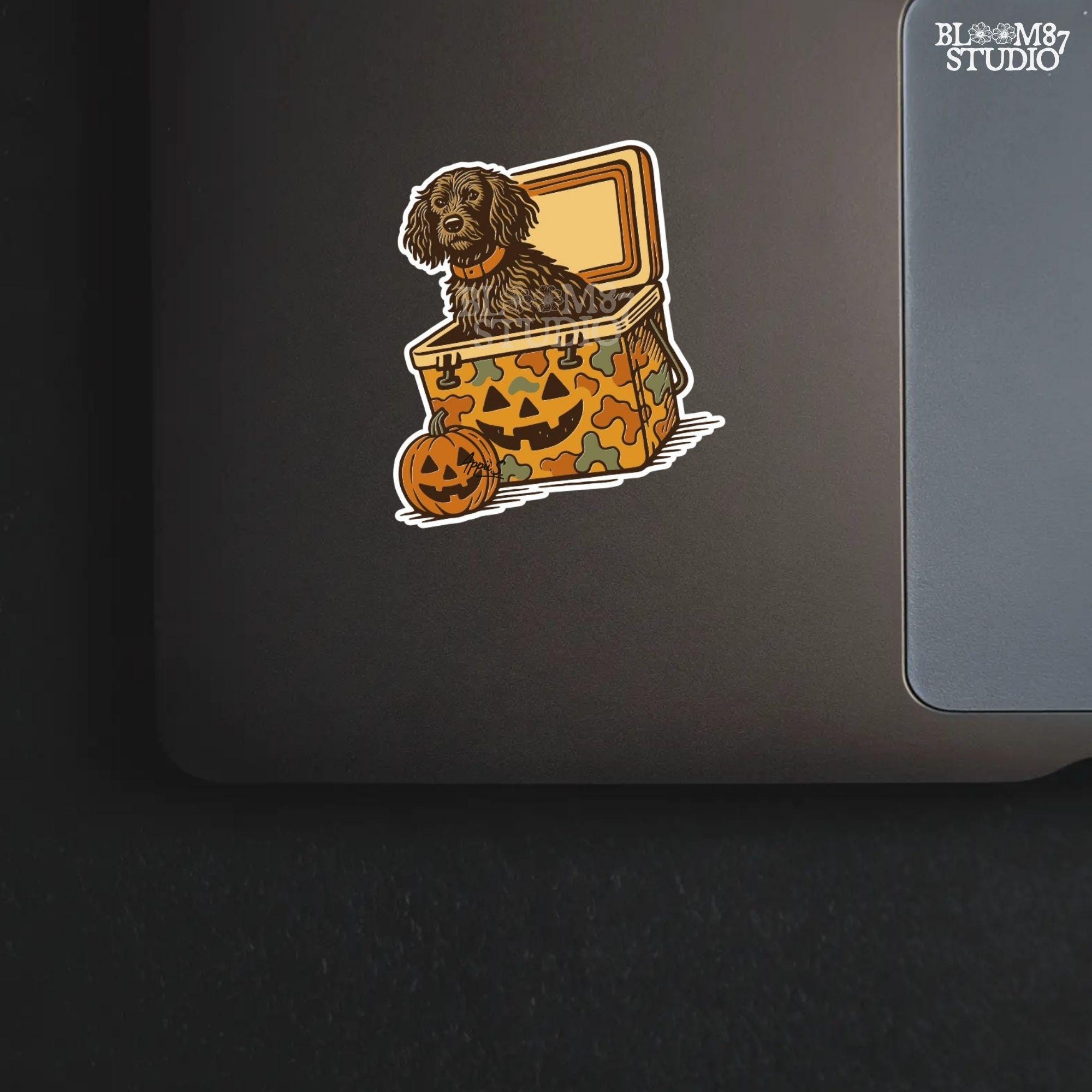 Halloween-themed PNG of a fluffy dog sitting inside a pumpkin-faced camouflage cooler, with jack-o’-lantern pumpkins next to it.