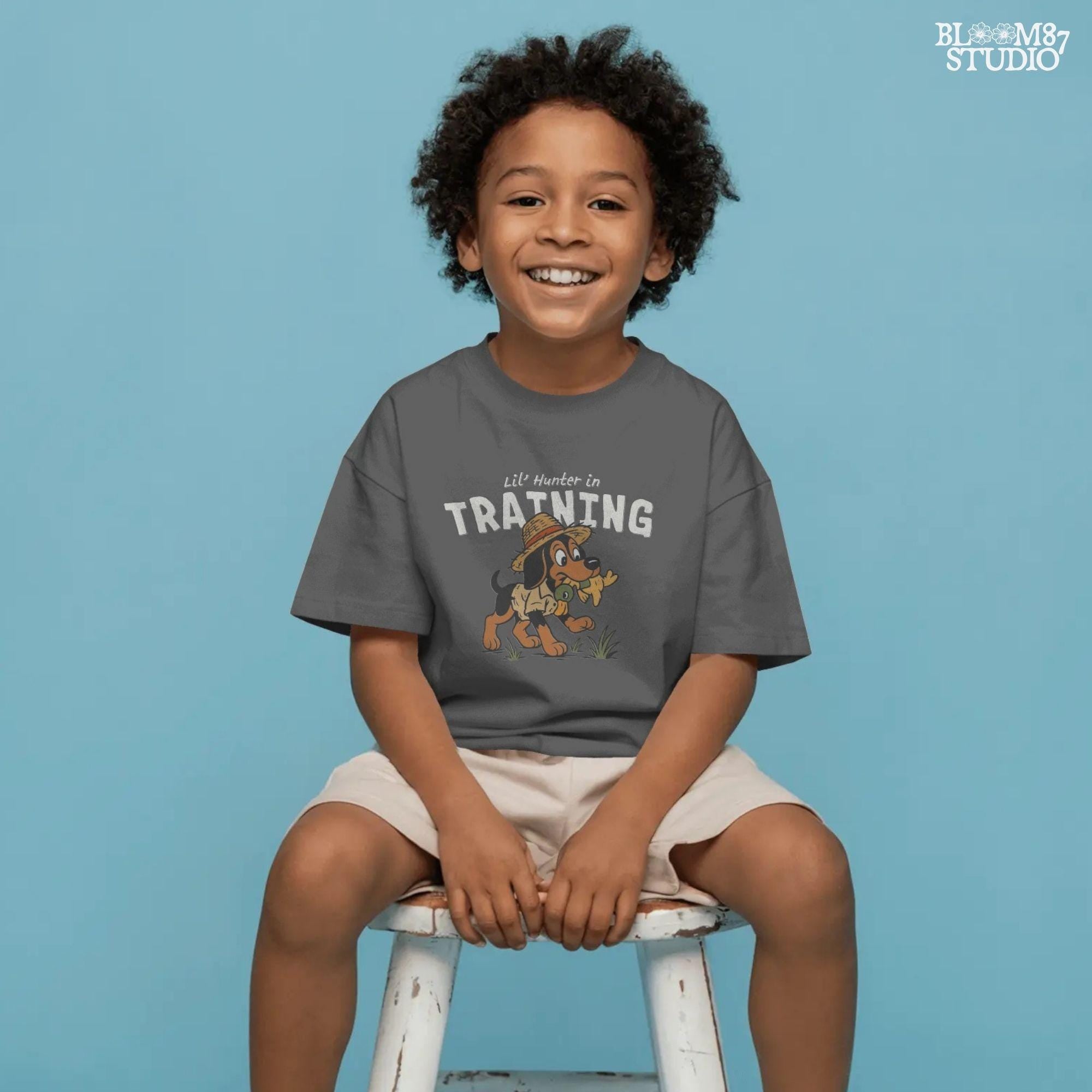 Cartoon hunting dog wearing straw hat and camo shirt holding duck toy in mouth with “Lil’ Hunter in Training” text – cute PNG for kids sublimation.