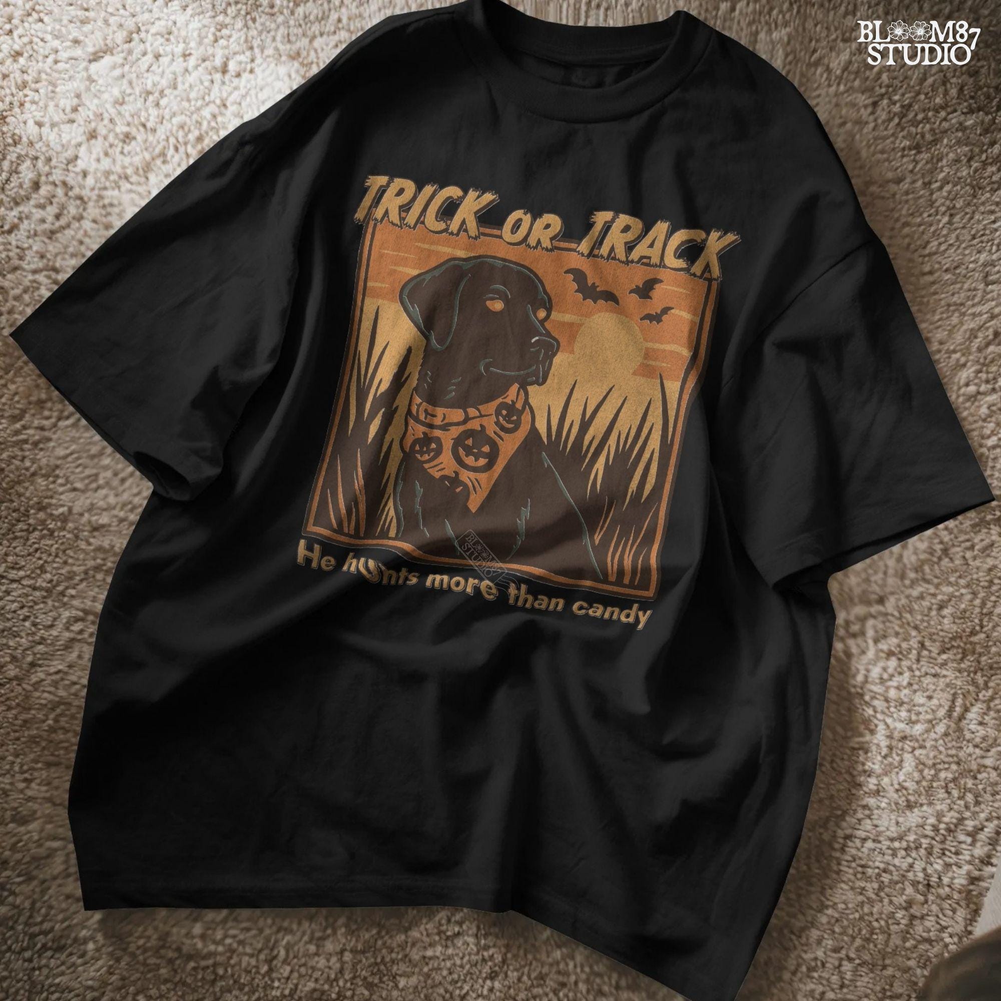 Halloween PNG of a black lab hunting dog wearing a pumpkin bandana with bats, sunset, and text “Trick or Track – He Hunts More Than Candy”