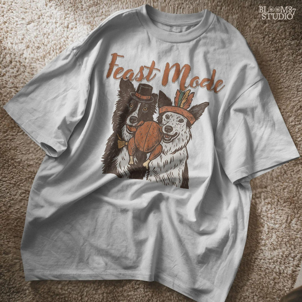 Rustic-style Thanksgiving dog clipart with two collies in pilgrim and feather costumes holding roasted turkey under Feast Mode text. Perfect for funny Thanksgiving shirts, mugs, fall crafts, and wall prints.